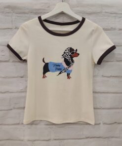 Camiseta de chica en tono crema con cuello redondo marrón, manga corta y estampado central de perro salchicha con pañuelo y l